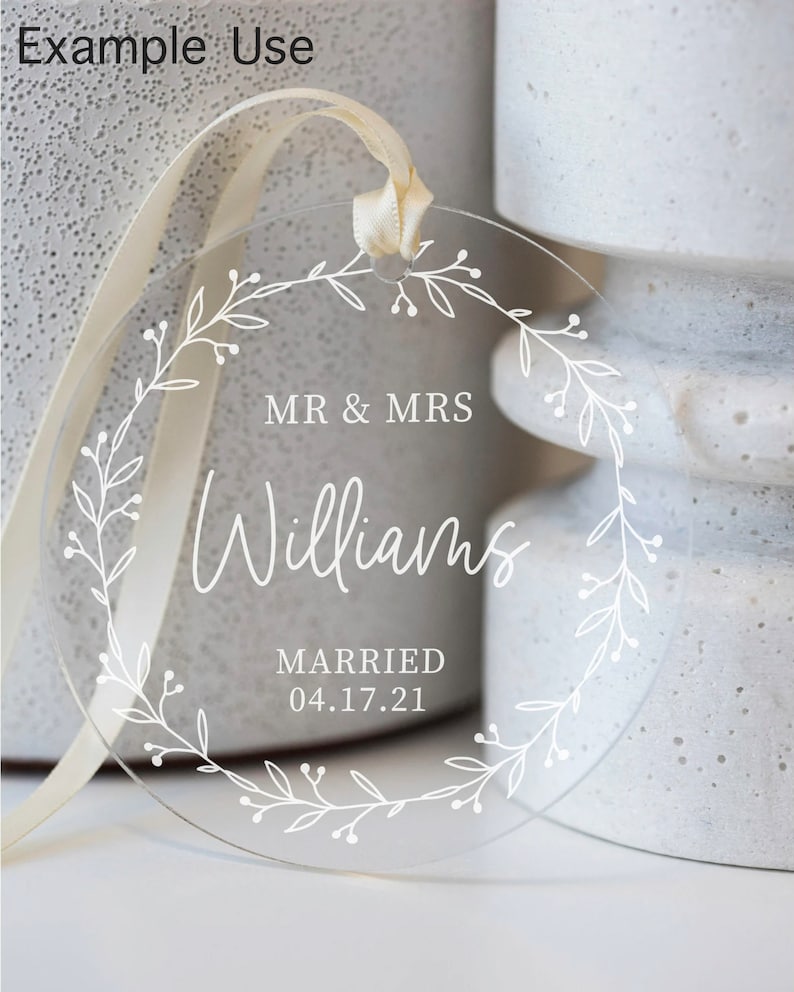 Puede incluir: Un adorno redondo de acr&iacute;lico transparente con un dise&ntilde;o de corona floral blanca. El adorno presenta el texto "MR & MRS Williams MARRIED 04.17.21".