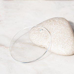 Puede incluir: Un c&iacute;rculo de vidrio transparente, o disco, sobre una superficie de m&aacute;rmol blanco. El c&iacute;rculo est&aacute; acostado de lado, con una piedra de color marr&oacute;n claro en forma de coraz&oacute;n detr&aacute;s.