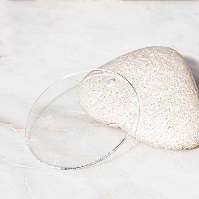 Puede incluir: Un c&iacute;rculo de vidrio transparente, o disco, sobre una superficie de m&aacute;rmol blanco. El c&iacute;rculo est&aacute; acostado de lado, con una piedra de color marr&oacute;n claro en forma de coraz&oacute;n detr&aacute;s.