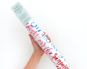 May include: A white table runner with blue and red watercolour lettering that says "Oh the places you'll go" and "Right on the ramparts". The table runner is rolled up and packaged in a clear plastic sleeve.