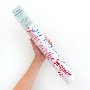 May include: A white table runner with blue and red watercolor lettering that says "Oh the places you'll go" and "Right on the ramparts". The table runner is rolled up and packaged in a clear plastic sleeve.