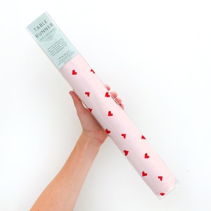 May include: Pink table runner with a red heart pattern. The runner is rolled up and packaged in a clear plastic sleeve. The label on the sleeve reads "Table Runner 18.5" x 54" Gift Wrap Paper".