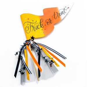 May include: A Halloween trick or treat wand with an orange, yellow, and white candy corn design. The wand has black and white ribbon streamers and black star-shaped embellishments.
