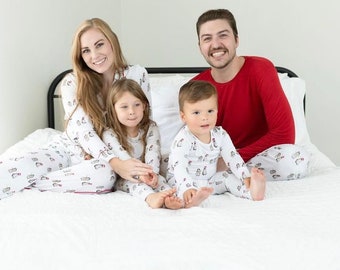 May include: A family of four wearing matching white pajamas with a penguin design. The pajamas have red trim and a red Santa hat on the penguin. The family is sitting on a white bed with a white pillow behind them. The father is wearing a red long-sleeved shirt.