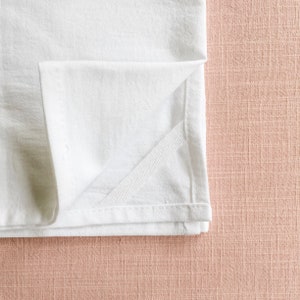 May include: A white kitchen towel folded in half, with a visible seam along the edge. The towel is on a light pink textured surface.