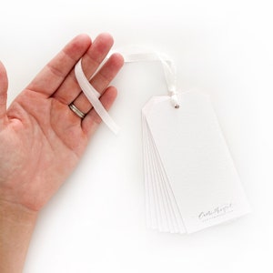 May include: A stack of white gift tags with a pink border and a silver eyelet. The tags are tied together with a white ribbon and have the text "Cathrynel" printed on the bottom.