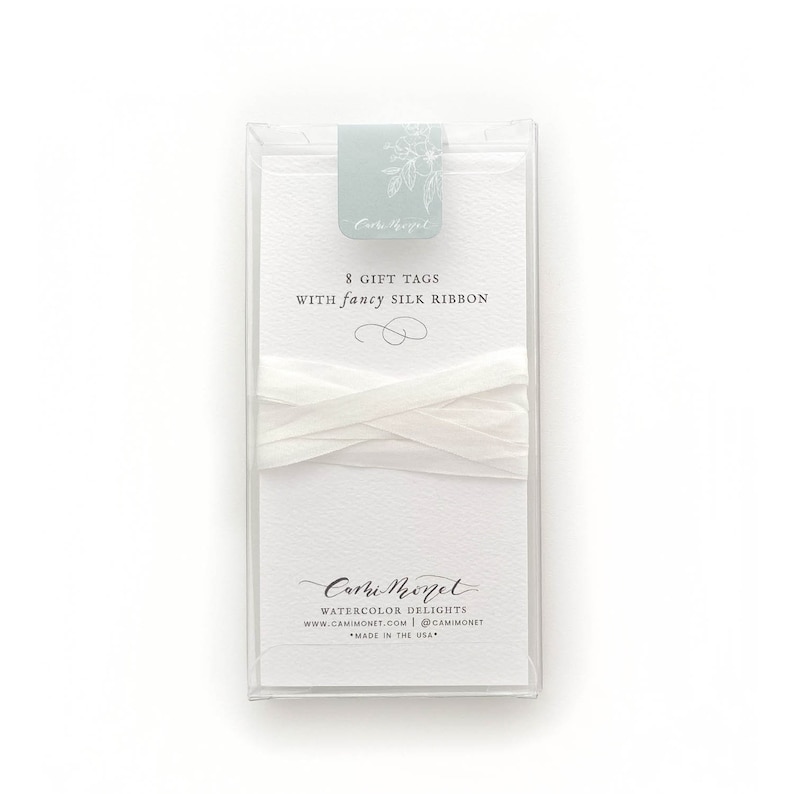 May include: A set of 8 white gift tags with a white silk ribbon. The tags are packaged in a clear plastic box with a blue and white label that says "Cami Monet Watercolor Delights". The label also includes the website address and social media handle for the company.
