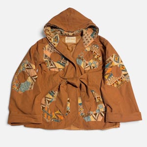 May include: A brown hooded jacket featuring a tie-front closure and large, hexagonal patches on the sleeves and front panels. The patches display a multi-colored geometric pattern in shades of blue, orange, and cream. A label reads MOD-MAID.