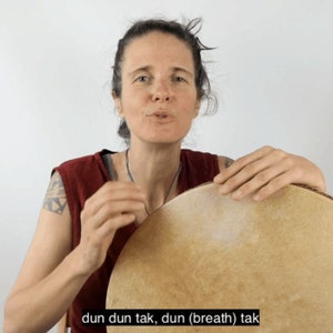 Peut inclure: Une femme tient un grand tambour rond avec une surface brun clair. Le texte "dun dun tak, dun (breath) tak" est affiché en bas de l'image. La femme porte un haut rouge et a un tatouage sur le bras.