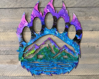 May include: A wooden bear paw print with a mountain and lake scene painted in shades of blue, green, purple, and pink.
