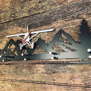 May include: Black metal wall hook shaped like a mountain range with a small airplane flying over it. The hook has eight hooks for hanging items.