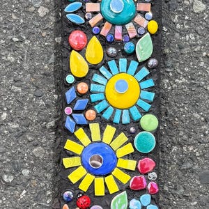 May include: A rectangular mosaic wind chime with a black background. The mosaic features colorful flower designs in yellow, blue, orange, and pink. A small silver bell hangs at the bottom. The mosaic is made of glass and ceramic.
