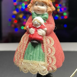 May include: A ceramic Christmas ornament depicting a girl in a red coat holding a smaller doll. The ornament has a green dress, a green bow, and a lace trim. The background is blurred with colorful lights.