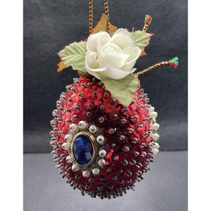 May include: A red sequined ornament with a white flower and green leaves. The ornament features a blue jewel surrounded by faux pearls. A decorative holiday item.