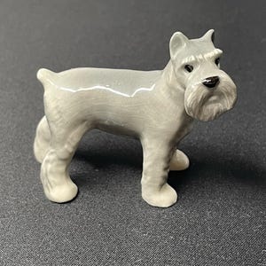 May include: A ceramic figurine of a Schnauzer dog in a standing pose. The dog is primarily light gray with a white beard and chest. The figurine has a glossy finish and is set against a dark gray background.