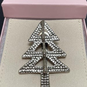 May include: Silver-toned Christmas tree brooch with a textured, dotted surface. The brooch is displayed inside a pink gift box with a light beige interior. The tree design features a vertical bar and a textured surface.