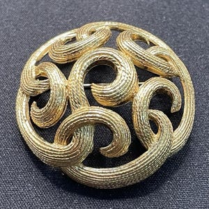 以下が含まれることがあります： 円形のデザインの金色のブローチ。ブローチは、円形のフレーム内に、相互接続された、テクスチャ加工された、渦巻く形状のシリーズを備えています。 テクスチャ加工された表面が光を捉え、ハイライトと影を作り出します。 ブローチは暗い背景に配置されています。