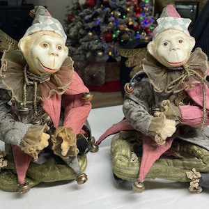 May include: Two decorative monkey figurines dressed as jesters. Each has a white face, a checkered hat, and a ruffled collar. The jesters wear pink and gray outfits with gold accents and sit on green cushions.