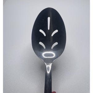 Puede incluir: Una cuchara ranurada de acero inoxidable con cabeza ovalada y mango largo. La cuchara presenta una serie de formas recortadas decorativas en la cabeza. La cuchara está diseñada para escurrir líquidos al servir.