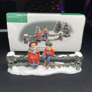Op de afbeelding: Een Department 56 Village Accessories Split Rail Fence beeldje. Het beeldje toont twee kinderen in winterkleding die op een met sneeuw bedekte hek zitten, versierd met hulst. De doos toont een vergelijkbare scène. De kinderen dragen rode jassen, hoeden en sjaals.