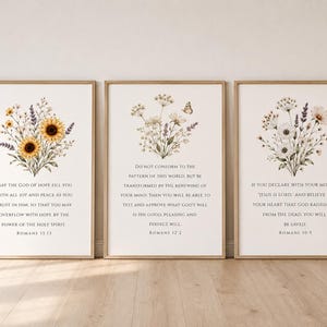 May include: Three framed wall art prints featuring floral bouquets and Bible verses. The left print shows sunflowers and Romans 15:13. The center print displays wildflowers, a butterfly, and Romans 12:2. The right print features white flowers and Romans 10:9.