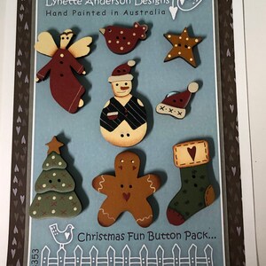 May include: A set of nine hand-painted wooden Christmas buttons featuring a snowman, gingerbread man, angel, bird, star, stocking, and Christmas tree. The buttons are on a blue background with a white picket fence and the text "Christmas Fun Button Pack..."