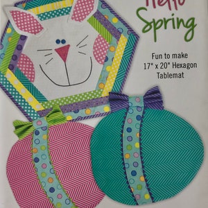 May include: A sewing pattern for a set of spring-themed table linens, including a 17-inch by 20-inch hexagon table mat, a 15-inch by 16-inch egg placemat, and a napkin. The pattern features a bunny, a bow, and eggs with colorful polka dots and stripes.
