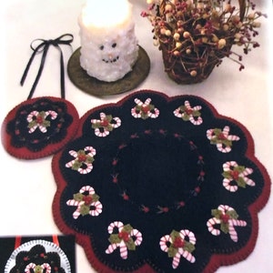 May include: A set of three felt Christmas ornaments with a candy cane and holly design. The ornaments are in shades of red, green, white, and black. The largest ornament is a round table topper, and the two smaller ornaments are round coasters.
