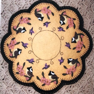 May include: A round, cream-colored felt rug with a black border. The rug is decorated with a circle in the center and a ring of embroidered birds, butterflies, and pink flowers around the circle.