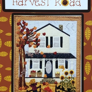 May include: A quilted wall hanging with a white house, a brown tree, and a scarecrow in front of it. The house has a blue door and a porch with a white railing. The tree has red and orange leaves. The scarecrow is wearing a brown hat and a brown shirt. The text "Harvest Road" is at the top of the image.