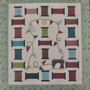 May include: A quilt block with a sewing theme. It features a variety of spools of thread in different colors and patterns, a pair of scissors, and a vintage sewing machine. The spools are arranged in a grid pattern, with a vine of hearts and leaves winding around them.