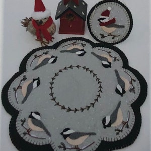 May include: A gray felt rug with a black border and a wreath of brown leaves in the center. Eight black-capped chickadees are embroidered on the rug, each with a white breast and a black cap. The rug is surrounded by a red birdhouse, a gray felt ornament with a chickadee, and a gray felt owl with a red Santa hat.