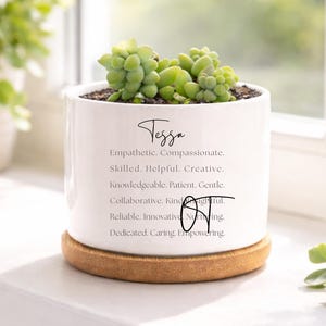 May include: A white ceramic planter with the name "Tessa" and inspirational words printed on it, holding a green succulent plant. The planter sits on a round, light brown cork coaster, placed on a white windowsill.