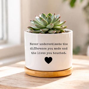 Puede incluir: Una maceta de cerámica blanca con una suculenta, sobre un posavasos de madera. La maceta tiene el texto "Never underestimate the difference you made and the lives you touched" y un corazón negro.