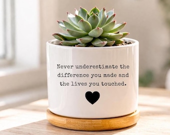 Può includere: Un vaso di ceramica bianco con una pianta succulenta, su un sottobicchiere di legno. Il vaso ha il testo "Never underestimate the difference you made and the lives you touched" e un cuore nero.
