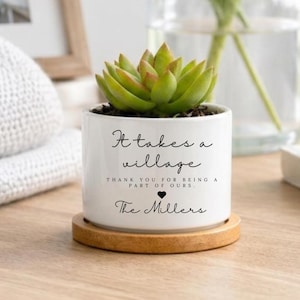 May include: A white ceramic succulent planter with a bamboo saucer. The planter features black text that reads: It takes a village, thank you for being a part of ours, with a small heart icon and the name The Millers printed below the text.