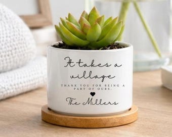 May include: A white ceramic succulent planter with a bamboo saucer. The planter features black text that reads: It takes a village, thank you for being a part of ours, with a small heart icon and the name The Millers printed below the text.