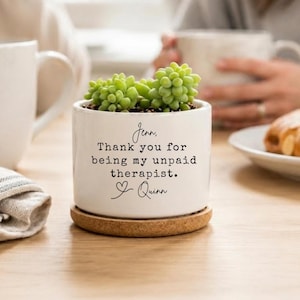 May include: A white ceramic cylindrical planter with a cork base containing a small green succulent. The planter features black printed text that reads: Jenn, Thank you for being my unpaid therapist. Quinn, with a small heart icon below the name.