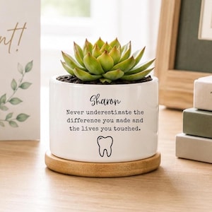 Puede incluir: Una maceta cilíndrica de cerámica blanca sobre una base de madera circular que contiene una suculenta verde. La maceta presenta el texto "Sharon Never underestimate the difference you made and the lives you touched." y un pequeño dibujo lineal de un diente.