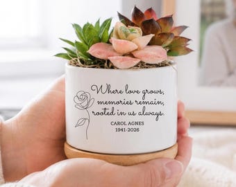May include: A white ceramic planter with a wooden base containing a succulent plant. The planter features a black line drawing of a rose and the text: Where love grows, memories remain, rooted in us always. CAROL AGNES 1941-2026.