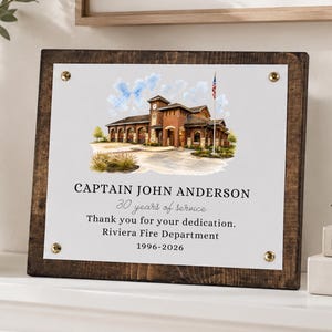 Puede incluir: Una placa rectangular de madera con un panel central blanco que presenta una ilustraci&oacute;n en acuarela de una estaci&oacute;n de bomberos y el texto: CAPTAIN JOHN ANDERSON, 30 years of service, Thank you for your dedication, Riviera Fire Department, 1996-2026.