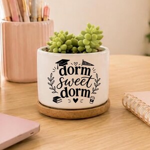 May include: A white ceramic planter with the text "dorm sweet dorm" in black script, featuring a graduation cap, books, and a coffee cup. The planter holds a green succulent and sits on a round, cork coaster. A pink pencil holder is in the background.