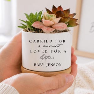 May include: A white ceramic planter with a wooden base containing a variety of succulents. The planter features the black printed text: CARRIED FOR A moment LOVED FOR A lifetime BABY JENSON. The design is simple, clean, and cylindrical in shape.