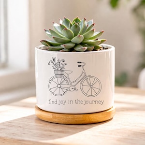 以下が含まれることがあります： 黒い自転車のイラストと「find joy in the journey」の文字が入った白いセラミックプランター。プランターには緑色の多肉植物が入っています。丸い木製のベースの上にあり、観葉植物に最適です。