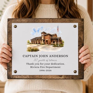 Puede incluir: Una placa rectangular de madera con un panel central blanco que presenta una ilustraci&oacute;n en acuarela de una estaci&oacute;n de bomberos y una bandera estadounidense. El texto dice: CAPTAIN JOHN ANDERSON, 30 years of service, Thank you for your dedication, Riviera Fire Department, 1996-2026.