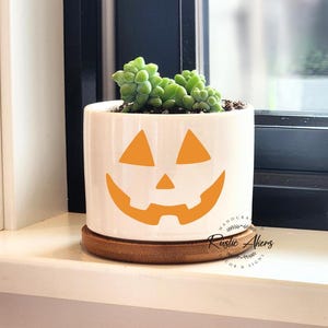 May include: White ceramic planter with a jack-o'-lantern face design. The planter has a brown wooden base and is filled with green succulents.