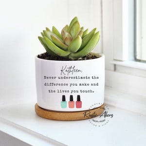 May include: White ceramic planter with a succulent plant and the text "Kathleen. Never underestimate the difference you make and the lives you touch."  Three nail polish bottles are depicted below the text. The planter has a wooden base.
