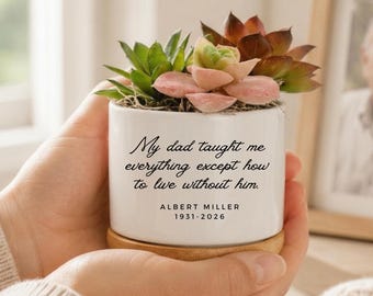 May include: A white ceramic cylindrical planter with a wooden base, containing various green and pink succulents. The front features black text: My dad taught me everything except how to live without him. ALBERT MILLER 1931-2026.