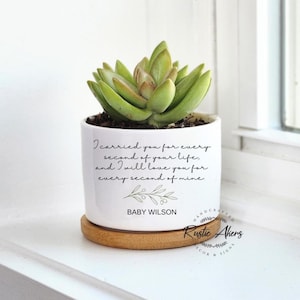Puede incluir: Una maceta de cerámica blanca con una planta suculenta. La maceta tiene una base de madera y presenta el texto "I carried you for every second of your life, and I will love you for every second of mine." y "BABY WILSON."
