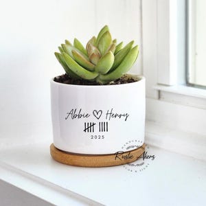 Puede incluir: Una maceta de cerámica blanca con una planta suculenta, sobre un platillo de madera. La maceta tiene los nombres "Abbie & Henry" y el año 2025 impresos. La maceta está en un alféizar blanco.